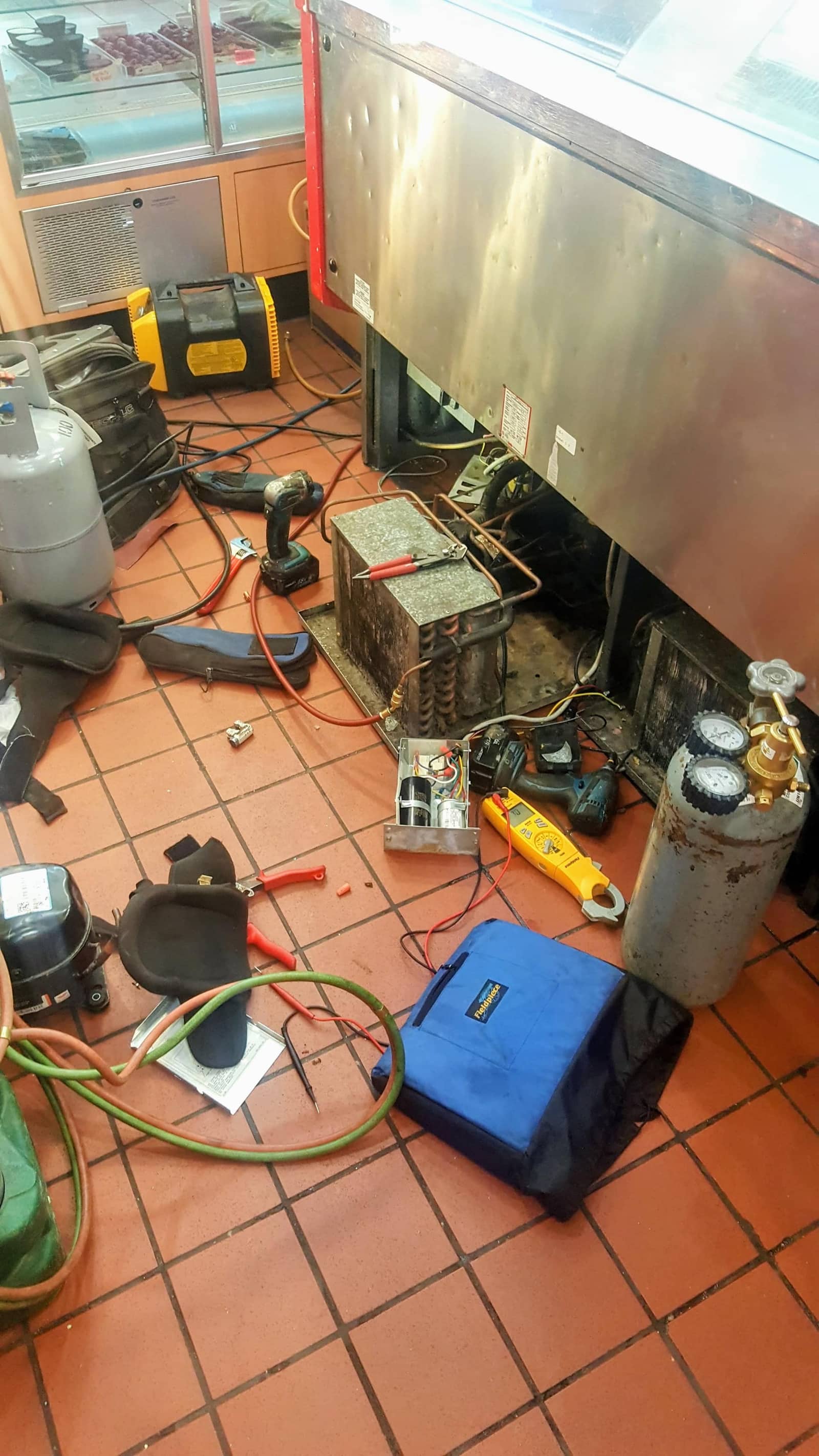 Technician servicing a commercial refrigeration display case with tools and gauges laid out
